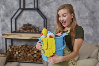 A cleaning woman is standing in living room holding a blue bucket fulfilled with chemicals and