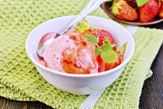 Strawberry ice cream in a white bowl with strawberries, strawberry syrup and a spoon on a napkin on