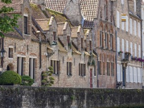 Row of cosy-looking, brick-clad historic buildings on a canal bank with lush greenery, Bruges,