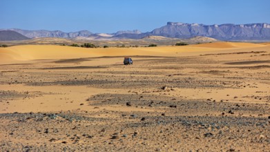 An off-road vehicle moves through a vast landscape of sand dunes and mountains, With off-road