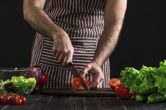 Man in a striped apron preparing salad on a wooden table. Men's hands cut the tomato to make a