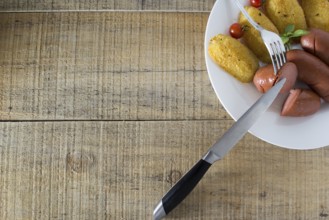 Pork sausages and breaded potatoes fried in a dish and decorated with cherry tomato. Fork and knife