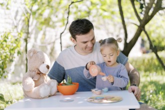 Little girl pours tea from a toy teapot into a toy cup while sitting on her dad lap at the table.