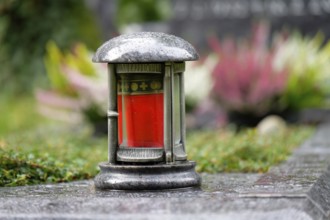 A grave lantern stands on the stone border of a grave with flowers in the blurred background