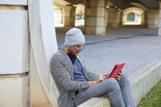Modern young man with tablet pc touch working under a bridge with wool winter cap