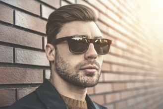 Portrait of stylish handsome young man with bristle standing outdoors and leaning on brick wall.