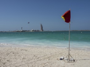 A red warning sign stands on the beach with turquoise water and a blue sky in the background,