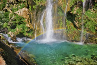 Waterfall with rainbow, surrounded by rocks and green foliage under a clear sky, Topoljski Buk