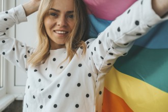 Young blonde millennial hippie woman with rainbow LGBTQ flag at home balcony. Peace and freedom the