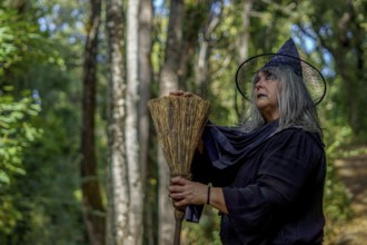 Old witch in black, dressed in black, seen in profile with her broom with a forest in the