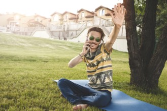 Young stylish handsome man is sitting on grass, resting and talking on the phone. waves his hand