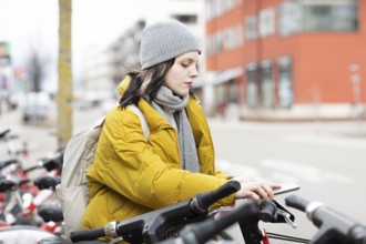 Woman in yellow jacket and cap on rental bikes in city street, cold winter day, environmentally