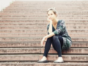 Sad lonely young woman sitting on steps. Portrait of serious girl with facial expression outdoors