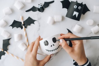 Female hands paint on white cup scary jacks face DIY for kids Halloween home activities Holiday art