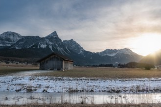 Winter alpine landscape with a wooden cabin, a frozen river and the snowy Alps mountains at sunset,