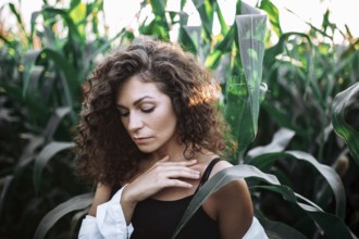 A serene woman with curly hair touches her chest in a sunlit cornfield