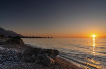 A peaceful sunset over the ocean with silhouette of mountains and pebble beach iwth rock boulders