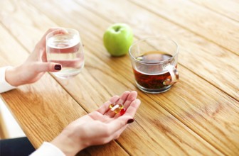 High angle of anonymous female with pills and cup of water witting at table with tea and apple