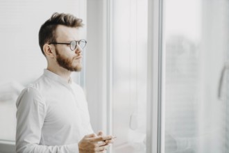 Man in glasses and white shirt holding a phone, looking thoughtfully out the window
