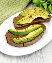 Two slices of rye bread with slices of avocado and pepper on a plate, napkin, parsley on the