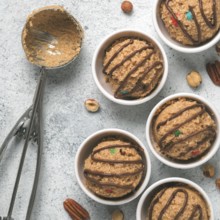 Safe-to-eat raw monster cookie dough in small portion bowl, ice cream scoop and nuts on gray cement