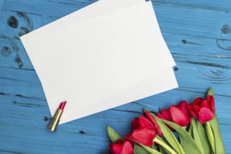 Red tulips, lipstick and a white sheet of paper for your greetings on the background of blue wooden