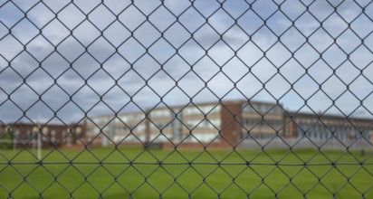A Typical High School Building And Sports Field Under A Cloudy Sky With Focus On The Security Fence