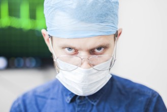 A surgeon with an intense expression, wearing mask and glasses, in a blurred clinical background