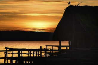 Amazing sunset landscape with a shadowed wooden hut and a bird on top of it, on a lake, at golden