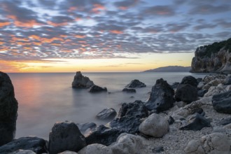 A colorful sunrise at Cala Gonone beach with black and white rocks and boulders in the foreground