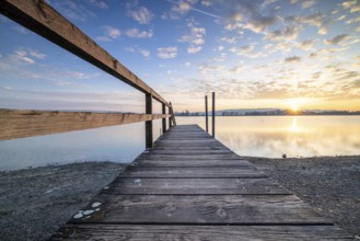 A wooden footbridge leading onto a calm lake as the sun sets under a cloudy sky, Naturfreundehaus,