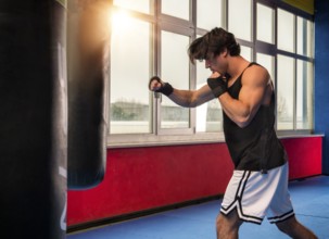 Side view of muscular sportive man in modern gym training with punching bag while boxing alone in