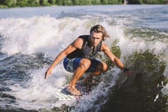 Beautiful long haired blond wakesurf in vest on board along waves of lake. Athletic male athlete