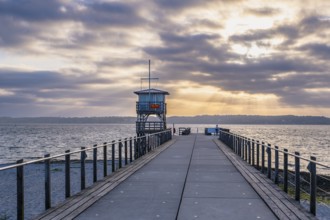 A long jetty with a tower leading into the sea under a dramatically cloudy sky with rays of