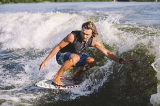 Young athletic man with long hair wakesurfing on waves of river in sunny summer weather. Ttheme