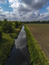 A quiet river runs between a planted field and a row of trees under a cloudy sky, aerial view of a