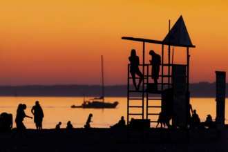 Silhouetted figures against an orange sunset on the beach with a lifeguard tower and a calm Lake