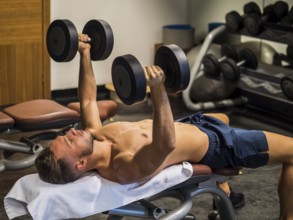 Muscular young man shirtless, lifting dumbbells training pecs on gym bench