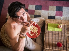 From above shot of happy muscular shirtless man sitting on floor having breakfast eating cereal,