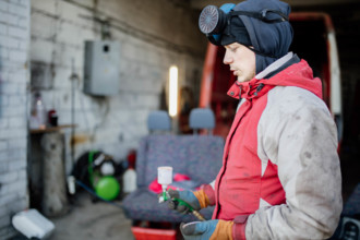 A repairman in a red jacket holds equipment, pausing work in a busy garage setting