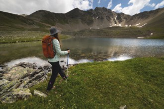 A woman with an orange backpack and a hat goes to a transparent lake in the mountains on a clear