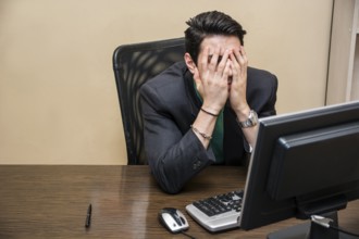 Desperate, worried young businessman sitting at his desk in front of his computer with his face in