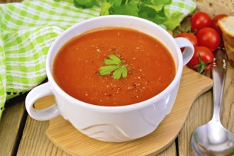 Tomato soup in a white bowl, green napkin, spoon, tomato, parsley, bread on a wooden boards