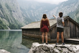 Two girls standing on top of a rock and wooden cottage in beautiful lake in the Alps with misty