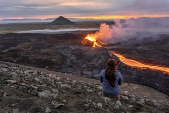 A woman sits on a mountain and looks at a glowing volcano, Grindavik, Sudurnes, Iceland, Europe