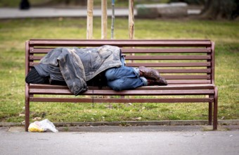 Homeless man sleeps on a bench covered with his jacket in a public park during the day