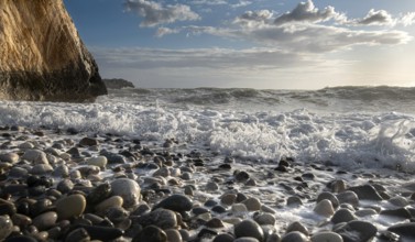 Low angle view of seascape with windy waves splashing on the rock and the sea pebbles. Rock of