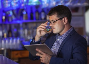 A presentable man in a bar, making a sip from a glass and meanwhile watching at his pad