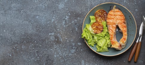 Grilled fish salmon steak and green salad with lemon on ceramic plate on rustic blue stone