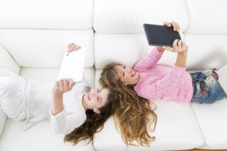 Children friends kid girls having fun playing with tablet pc lying on white sofa